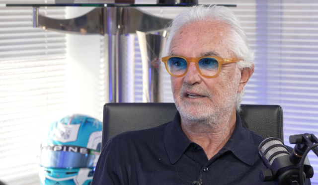 Older man with white hair and orange glasses speaks into a microphone in a studio.