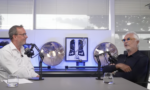 Two men in a podcast studio sit across from each other with microphones, glasses of water, and sports memorabilia in the background.