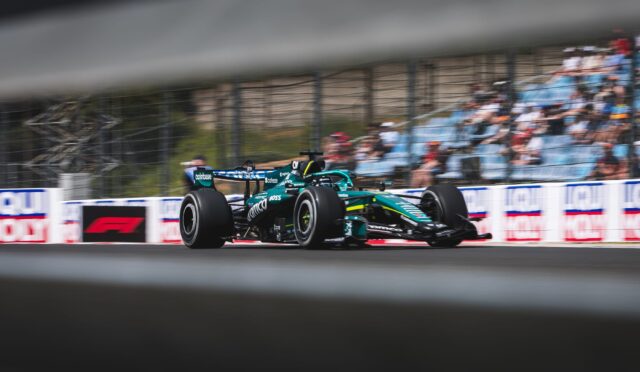 Formula 1 car racing at speed on a track, with a blurred grandstand crowd in the background and motion blur on the wheels.