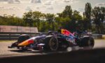 Two Formula 1 cars racing side by side on a track, red bull livery visible, with safety barriers and trees in the background.