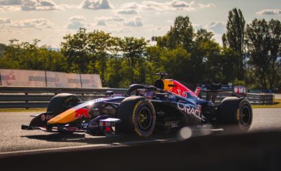 Two Formula 1 cars racing side by side on a track, red bull livery visible, with safety barriers and trees in the background.