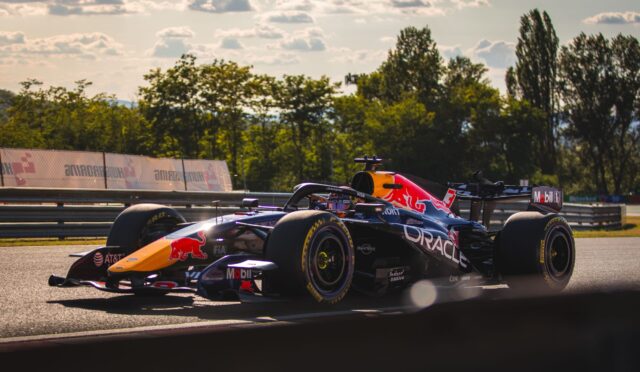 Two Formula 1 cars racing side by side on a track, red bull livery visible, with safety barriers and trees in the background.