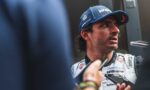 A race car driver in a Williams F1 uniform being interviewed, looking to the right, cap visible with blue branding.