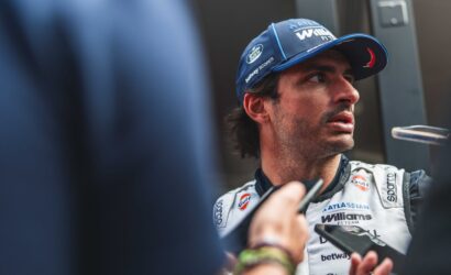 A race car driver in a Williams F1 uniform being interviewed, looking to the right, cap visible with blue branding.