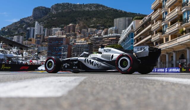 Formula 1 car on a street circuit with city buildings and a mountain backdrop in clear blue sky.