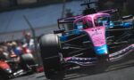 Front-on view of a pink-and-blue Formula 1 car speeding around a track, with blurred spectators in the background.