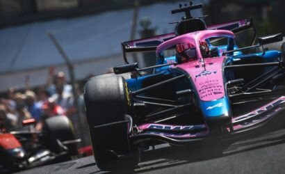Front-on view of a pink-and-blue Formula 1 car speeding around a track, with blurred spectators in the background.