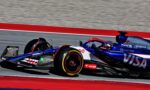 Formula 1 race car in blue/red livery speeds on a track with red and blue curbs, sponsor logos visible on the sidepod and rear wing.