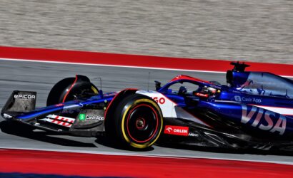 Formula 1 race car in blue/red livery speeds on a track with red and blue curbs, sponsor logos visible on the sidepod and rear wing.