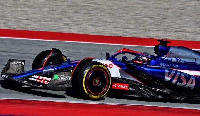 Formula 1 race car in blue/red livery speeds on a track with red and blue curbs, sponsor logos visible on the sidepod and rear wing.