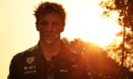 Male race car driver in a sponsor-covered racing suit during sunset, facing the camera.