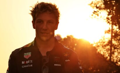 Male race car driver in a sponsor-covered racing suit during sunset, facing the camera.
