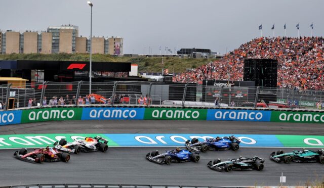 Formula 1 race in progress: several F1 cars cornering on a track with a packed grandstand and sponsor barriers along the wall.