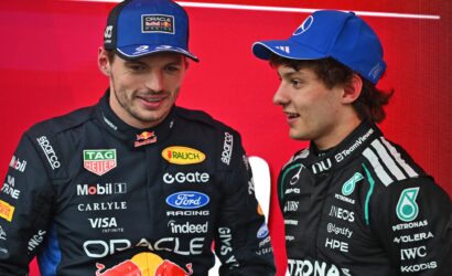 Two race car drivers in branded racing suits converse in front of a bright red backdrop, with sponsor logos visible on their suits.