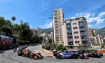 Formula 1 cars race around a sharp city corner, lined by high-rise buildings and green hills under a blue sky.