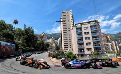Formula 1 cars race around a sharp city corner, lined by high-rise buildings and green hills under a blue sky.