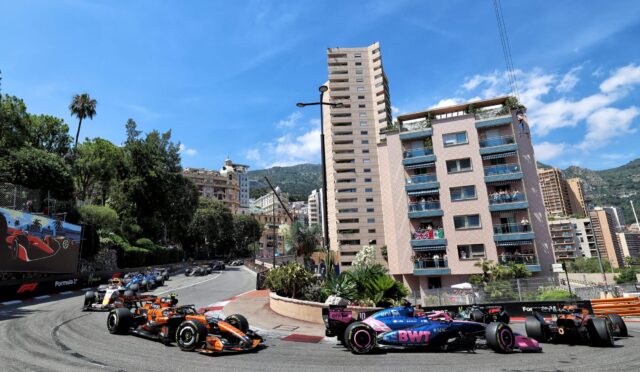 Formula 1 cars race around a sharp city corner, lined by high-rise buildings and green hills under a blue sky.