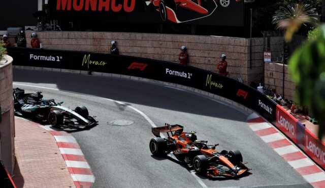 Formula 1 cars race along Monaco’s street circuit, tight barrier walls with sponsor banners (Formula 1, Monaco, Lenovo), spectators in red uniforms watch from the sides