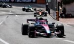 Pink and blue Formula 1 car leads a street circuit race, with other cars in the distance and barriers along the track behind it.