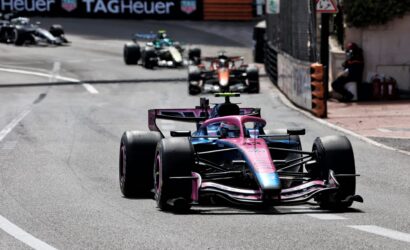 Pink and blue Formula 1 car leads a street circuit race, with other cars in the distance and barriers along the track behind it.