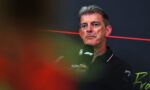 Man with gray hair speaking into a microphone at a Formula 1 press event, black polo with F1 logos, dark backdrop with a red line behind him.