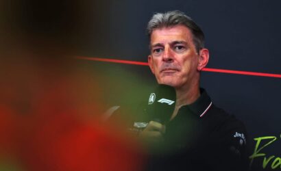 Man with gray hair speaking into a microphone at a Formula 1 press event, black polo with F1 logos, dark backdrop with a red line behind him.