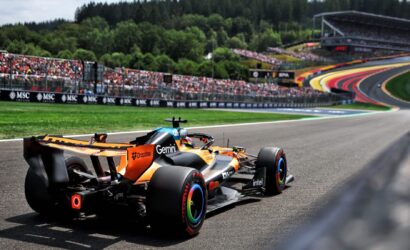 Orange Formula 1 car on a race track, crowds in the stands and trees in the background. View focuses on the car as it rounds a bend.