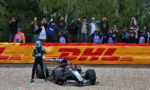 Formula 1 car on track with driver in blue helmet, crowd behind a fence taking photos. DHL banner in front.