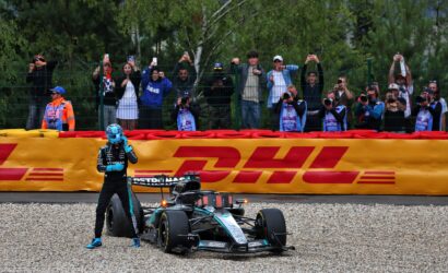 Formula 1 car on track with driver in blue helmet, crowd behind a fence taking photos. DHL banner in front.