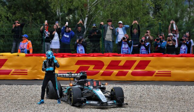 Formula 1 car on track with driver in blue helmet, crowd behind a fence taking photos. DHL banner in front.