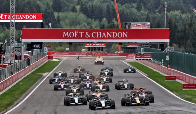 Starting grid of Formula 1 cars on a track, with Moët & Chandon branding overhead and green forest backdrop.