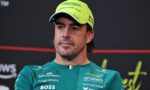 Man in a teal Aston Martin Formula One Team shirt wearing a neon yellow cap, looking off to the side at a press event in a dark background.