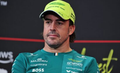 Man in a teal Aston Martin Formula One Team shirt wearing a neon yellow cap, looking off to the side at a press event in a dark background.