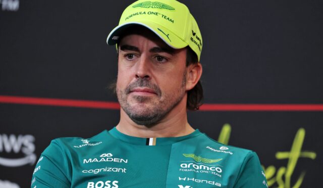 Man in a teal Aston Martin Formula One Team shirt wearing a neon yellow cap, looking off to the side at a press event in a dark background.