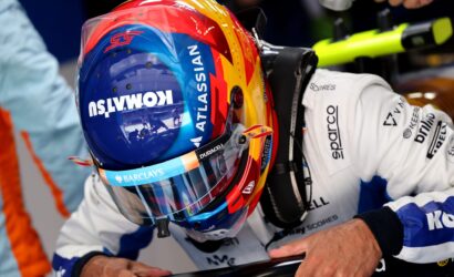 Racing driver in a colorful helmet and white suit, hands gripping a black race car barrier in a pit area