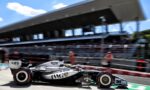 Motion-blurred Formula 1 car in the pit lane, black and silver livery with sponsor logos, grandstand and pit building in the background.