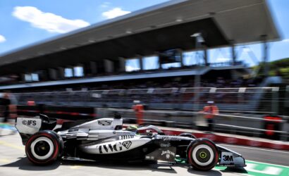 Motion-blurred Formula 1 car in the pit lane, black and silver livery with sponsor logos, grandstand and pit building in the background.