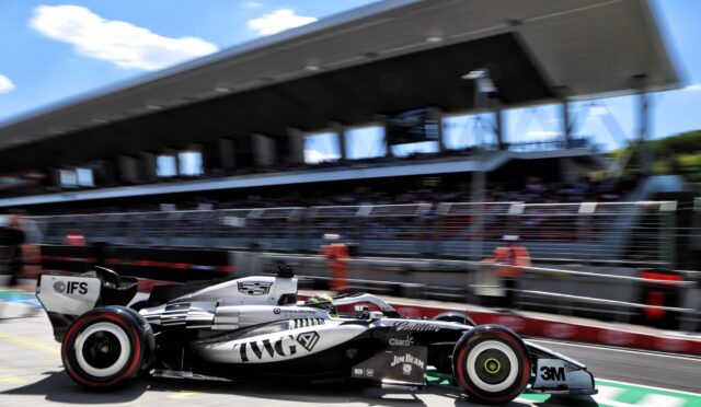 Motion-blurred Formula 1 car in the pit lane, black and silver livery with sponsor logos, grandstand and pit building in the background.