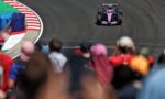 Pink and blue Formula 1 car on a dark race track, seen from amidst blurred spectators on the pit straight edge of the track barrier.