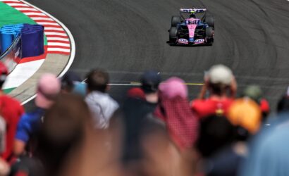 Pink and blue Formula 1 car on a dark race track, seen from amidst blurred spectators on the pit straight edge of the track barrier.