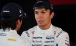 Race car driver in a white sponsor-labeled suit speaks with a teammate, wearing a blue cap with Kraken branding.
