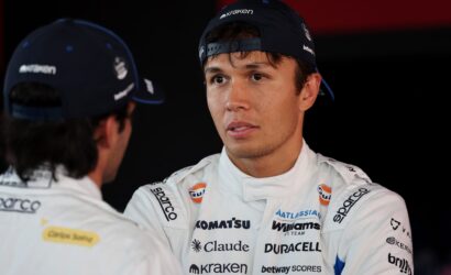 Race car driver in a white sponsor-labeled suit speaks with a teammate, wearing a blue cap with Kraken branding.