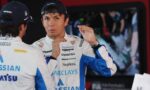 Race car driver in a white-blue suit with Barclays and Gulf logos talks to a teammate, wearing a navy cap and gesturing with his hands in the pit area.