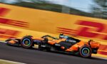 Orange Formula 1 race car speeding along a track with motion blur, sponsor logos visible on the side pod and wheels.