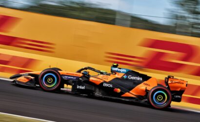 Orange Formula 1 race car speeding along a track with motion blur, sponsor logos visible on the side pod and wheels.