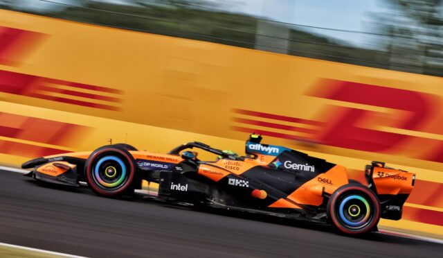 Orange Formula 1 race car speeding along a track with motion blur, sponsor logos visible on the side pod and wheels.