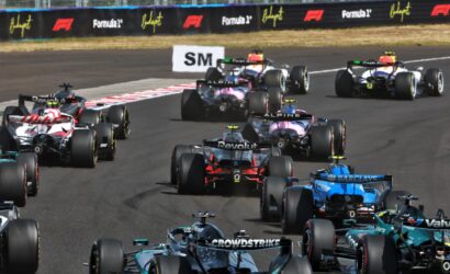 Formula 1 race cars clustered on a track, speeding past sponsor barriers in a tight pack.