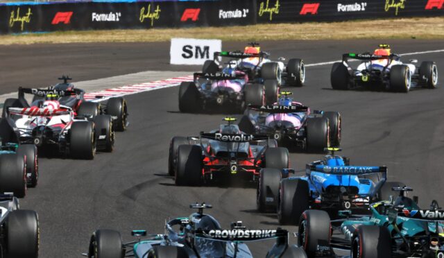 Formula 1 race cars clustered on a track, speeding past sponsor barriers in a tight pack.