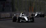 Two Formula 1 cars race on a sunlit track, closely following each other near the barriers with a bright green rear wing highlight on the lead car.