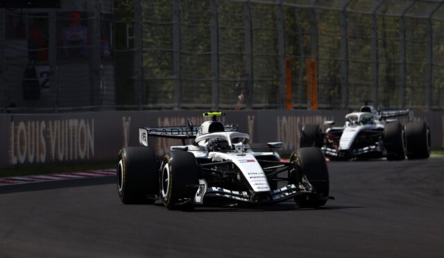 Two Formula 1 cars race on a sunlit track, closely following each other near the barriers with a bright green rear wing highlight on the lead car.